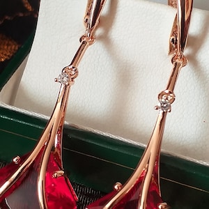 May include: A pair of rose gold earrings with a dangling design. Each earring features a red, faceted gemstone and a small, clear accent stone. The earrings are displayed in a green and white jewelry box.
