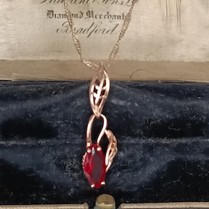 May include: A rose gold pendant necklace featuring a red gemstone. The pendant has an openwork design and a marquise-cut red stone. The necklace is displayed in a vintage jewellery box with the text "Falkiner & Sons Ltd Diamond Merchants Bradford".