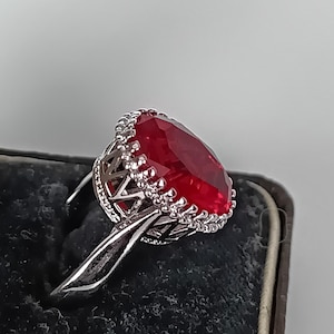 Può includere: Un anello color argento con una grande pietra preziosa rossa di forma ovale e sfaccettata. L'incastonatura presenta un bordo metallico decorativo smerlato attorno alla pietra e un design a griglia sui lati del gambo, che mette in mostra una lavorazione dettagliata.