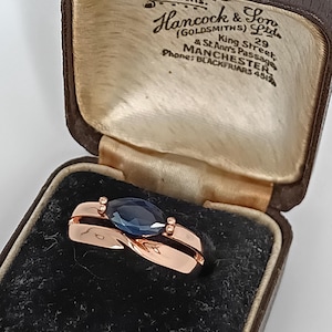 May include: A rose gold ring with a dark blue, oval-shaped gemstone, presented in a vintage brown jewelry box. The box's interior is lined with satin. The box has text that reads "Hancock & Son (Goldsmiths) Ltd".