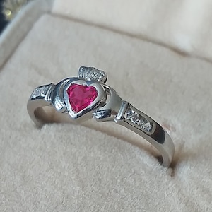 Peut inclure: Une bague celtique Claddagh en argent avec une pierre précieuse rouge en forme de cœur et de petites pierres précieuses claires sur la bande.