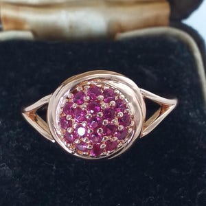 Op de afbeelding: Een roségouden ring met een cluster van kleine, ronde, helder roze edelstenen. De ring heeft een cirkelvormig midden met een band die aan elke kant in een driehoekig ontwerp splitst. De ring is geplaatst tegen een zwarte achtergrond.