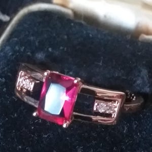 Könnte beinhalten: Ein Ring aus Roségold mit einem großen, quadratischen, roten Edelstein und kleineren, klaren Edelsteinen auf der Ringschiene.