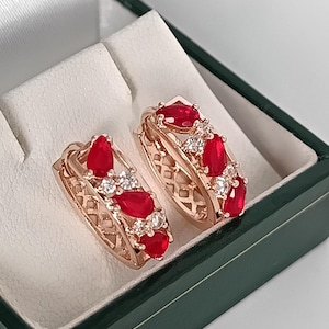 Puede incluir: Un par de pendientes de aro de oro rosa con gemas rojas en forma de lágrima y pequeñas gemas blancas. Los pendientes se exhiben en una caja de joyería verde con interior blanco. Los pendientes tienen un diseño intrincado.