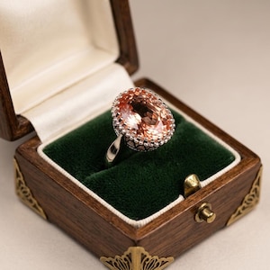 Puede incluir: Un anillo de plata con una gran piedra preciosa de corte ovalado en color melocotón, rodeada por un engaste de halo con cuentas. El anillo se presenta dentro de una caja de madera oscura con un forro de terciopelo verde y esquinas doradas.