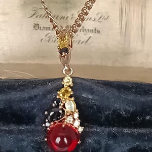 May include: A gold-toned necklace featuring a pendant with a large, round, deep red cabochon stone at the base. The pendant is accented with a cluster of smaller, faceted yellow, black, and clear stones arranged in an asymmetrical, organic shape.