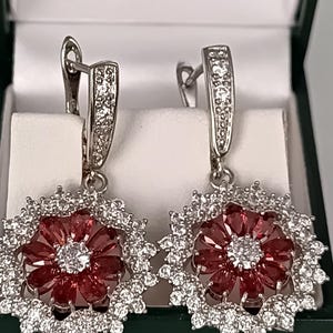Könnte beinhalten: Ein Paar silberfarbene Ohrringe mit floralem Design. Jeder Ohrring hat einen zentralen Cluster aus roten Edelsteinen, umgeben von einem Kranz aus klaren, funkelnden Steinen. Die Ohrringe werden in einer grünen Schmuckschatulle präsentiert.