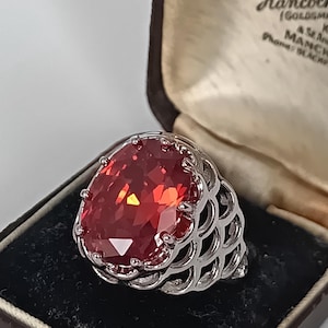 May include: A silver ring featuring a large, oval-cut, vibrant red gemstone held by multiple prongs. The band has an intricate, openwork lattice design. The ring is presented in a dark brown jewellery box with a cream-coloured interior lining.