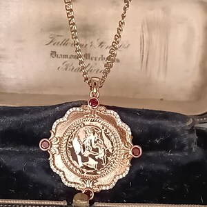 Op de afbeelding: Een goudkleurige hanger aan een ketting met een rond, muntachtig middendeel met een profielreliëf, omlijst door een geschulpte rand en geaccentueerd met vier kleine, ronde, donkerrode stenen op de windstreken, hangend aan een bijpassende schakelketting.