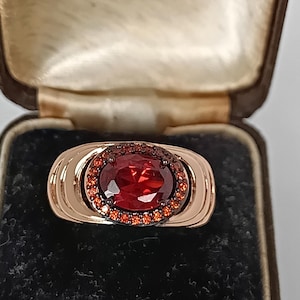 以下が含まれることがあります： 中央に大きな楕円形の深い赤色の宝石をあしらったローズゴールドのリング。周囲には小さなオレンジがかった赤色の石が配置されています。リングは長方形のデザインで、二重のバンドセッティングが施されています。ヴィンテージのジュエリーボックスに収められています。