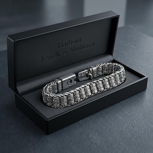 Pode incluir: Uma pulseira de metal prateado com uma série de elos cilíndricos com padrões em relevo intrincados, apresentada numa caixa retangular preta com o texto Harbour Jewellers, Mullingar gravado na tampa interior do estojo de joias.