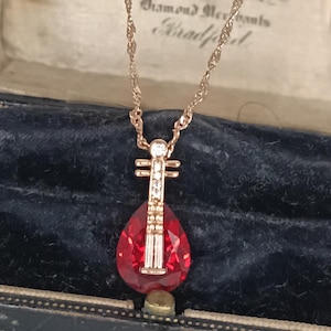 Op de afbeelding: Een goudkleurige ketting met een hanger in de vorm van een luit. De hanger heeft een grote, druppelvormige rode edelsteen en is versierd met kleine, heldere edelstenen. De ketting wordt gepresenteerd in een vintage sieradendoos.