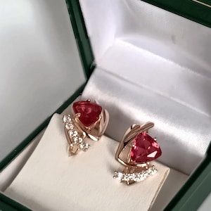 May include: A pair of rose gold earrings with a triangular, red gemstone center and small, clear accent stones. The earrings are displayed in a green jewelry box with a white satin interior.