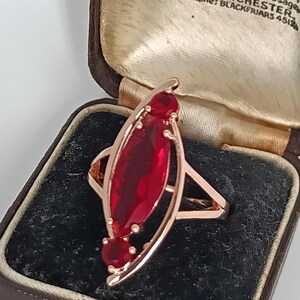 Peut inclure: Bague en or rose ornée d'une grande pierre rouge en forme de marquise et de deux petites pierres rouges. La bague est présentée dans un écrin à bijoux vintage marron. Le texte "Hancock & Son" est visible.