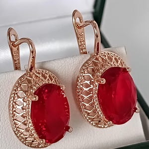 May include: Rose gold-tone earrings with large, oval, red gemstones. The earrings have a filigree design around the gemstones and a lever back closure. The earrings are displayed in a white box.
