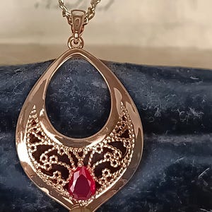 Op de afbeelding: Een roségouden hangerketting met een druppelvormig ontwerp. De hanger heeft een filigraan patroon en een felrode edelsteen in het midden. De ketting wordt gepresenteerd op een donkerblauwe ondergrond.