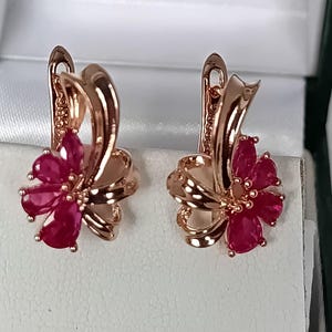 Könnte beinhalten: Ein Paar roségoldfarbene Ohrringe in Blumenform. Jeder Ohrring ist mit fünf birnenförmigen, tief rosa-roten Steinen besetzt, die wie Blütenblätter angeordnet sind und in einem polierten Metallrahmen mit sicherem Klappbügelverschluss sitzen.