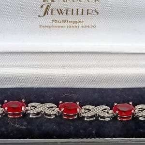 Op de afbeelding: Een zilverkleurige armband met ovaal geslepen rode stenen afgewisseld met X-vormige schakels bezet met kleine heldere steentjes. De armband zit in een doosje met de tekst Harbour Jewellers, Mullingar, Telephone: (044) 45470 aan de binnenzijde.