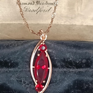 Op de afbeelding: Een goudkleurige hangerketting met een dieprode, marquise-vormige edelsteen. De hanger is omlijst door goudkleurig metaal en heeft twee kleinere rode edelstenen. De ketting wordt gepresenteerd in een vintage sieradendoos. De tekst "Saltorini & Sons Diamond Merchants Bradford" is zichtbaar.