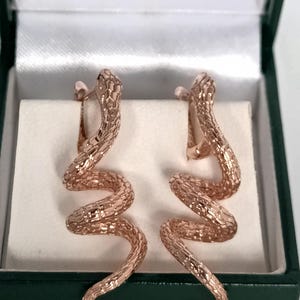 Op de afbeelding: Een paar roségouden slangen oorbellen in een groene sieradendoos. De oorbellen hebben een textuur met schubbenpatroon en een gebogen, serpentijnvormig ontwerp. Ze worden gepresenteerd op een wit, satijnachtig oppervlak.