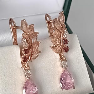 Op de afbeelding: Roségouden oorbellen met een bladvorm, met roze druppelvormige edelstenen. De oorbellen hebben ook kleine witte en rode accentstenen, gepresenteerd in een sieradendoosje.