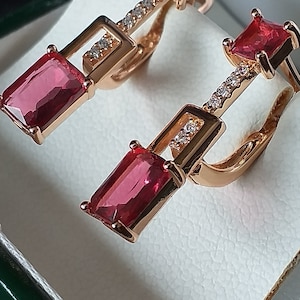 May include: A pair of rose gold earrings with red square-shaped gemstones and clear cubic zirconia stones. The earrings are in a small green box.