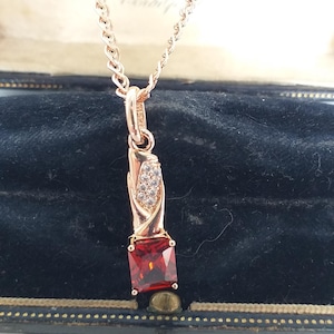 Könnte beinhalten: Halskette mit roségoldfarbenem Anhänger und einem rechteckigen, tiefroten Edelstein. Der Anhänger hat ein dekoratives Design mit kleinen, klaren Steinen. Die Halskette wird in einer Vintage-Schmuckschatulle mit schwarzem Samtinterieur präsentiert.