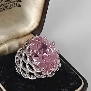 Può includere: Un anello color argento con una grande pietra preziosa rosa taglio ovale montata su griffe. La fascia dell'anello presenta un intricato design a reticolo traforato. L'anello è esposto all'interno di un portagioie marrone scuro con fodera color crema.