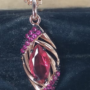 Könnte beinhalten: Halskette mit roségoldfarbenem Anhänger, der einen großen, tropfenförmigen roten Edelstein aufweist. Der Anhänger ist mit kleineren rosa und schwarzen Edelsteinen in einem blattartigen Design verziert. Die Kette ist ebenfalls roségoldfarben.