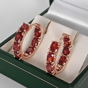 May include: Rose gold hoop earrings, each set with oval-cut, deep red gemstones. The earrings are presented in a green jewellery box with a white satin lining, highlighting their rich colour and design.
