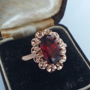 May include: A rose gold ring with a large, oval, deep red gemstone. The gemstone is surrounded by a scalloped, floral-like setting. The ring is displayed in a vintage jewelry box. The text "Hancock & Son" is visible.