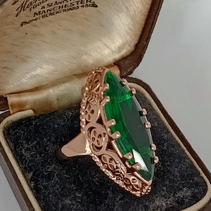 Puede incluir: Un anillo de oro rosa con una gran gema verde talla esmeralda. El anillo tiene un diseño de filigrana ornamentado alrededor de la gema. El anillo se muestra en un joyero vintage marrón.