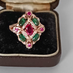 May include: Rose gold ring featuring a central oval ruby, encircled by green emeralds, small pink gems, and clear crystals. The ring has an intricate, swirling design and is presented in a brown jewellery box.