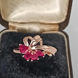 Könnte beinhalten: Ein Ring aus Roségold mit einer Anordnung von fünf birnenförmigen, tief rosa-roten Edelsteinen in Blumenform, akzentuiert durch ein poliertes, geschwungenes Metallbanddesign, das sich von der Basis der Steine entlang der Ringschiene erstreckt.