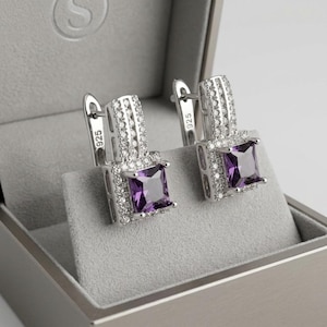 Op de afbeelding: Een paar oorbellen van sterling zilver met een vierkant geslepen paarse edelsteen omringd door kleine heldere steentjes. Het zilveren metaal is gestempeld met 925, wat duidt op een samenstelling van sterling zilver. Voorzien van een klapsluiting.