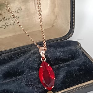 May include: A rose gold necklace with a large, faceted, teardrop-shaped red gemstone pendant. The pendant is suspended from a delicate chain. The necklace is displayed inside a vintage black jewelry box.