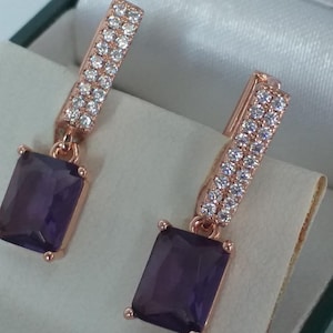 Op de afbeelding: Roségouden oorbellen met rechthoekige, dieppaarse edelsteenhangers. Het bovenste deel van de oorbellen is een staafje van kleine, heldere kristallen. De oorbellen worden gepresenteerd in een met witte satijn gevoerde doos.