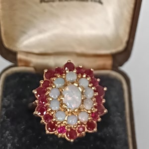 May include: A gold ring with a floral design, set with a central oval opal surrounded by small opals and red gemstones. The ring is displayed in a vintage jewellery box.