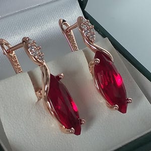 Può includere: Orecchini in oro rosa con una gemma rossa taglio marquise. Gli orecchini hanno un design curvo con piccole pietre trasparenti nella parte superiore. Presentati in un cofanetto verde scuro foderato di bianco.