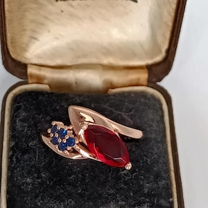 Könnte beinhalten: Ein roségoldener Ring mit einem großen, ovalen, roten Edelstein und einer Ansammlung kleiner blauer Edelsteine. Der Ring wird in einer Vintage-Schmuckschatulle präsentiert. Der Text "Hancock & Ltd" und "Manchester" ist sichtbar.