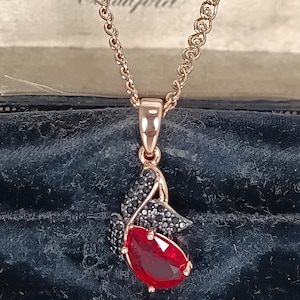 Könnte beinhalten: Halskette mit Anhänger aus Roségold, besetzt mit einem tropfenförmigen, facettierten roten Edelstein. Der Anhänger ist mit schwarzen Edelsteinen und einer zarten Roségoldkette verziert. Die Halskette wird vor einem dunkelblauen Hintergrund präsentiert.