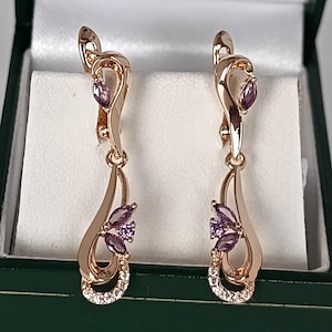 Peut inclure: Une paire de boucles d'oreilles en or rose avec un motif floral. Chaque boucle d'oreille présente une forme de goutte d'eau avec des accents de pierres précieuses violettes et un petit groupe de cristaux clairs. Les boucles d'oreilles sont dans un écrin vert.
