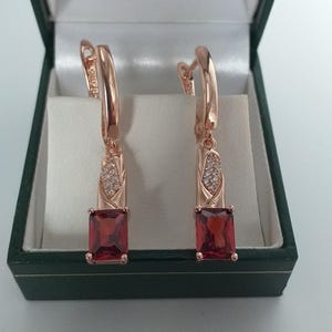 Può includere: Un paio di orecchini in oro rosa con una gemma rettangolare rosso scuro. Gli orecchini sono in un portagioie verde scuro con interno in raso bianco. Gli orecchini hanno una piccola sezione di pietre trasparenti.