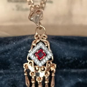 Puede incluir: Un collar con colgante dorado con una piedra roja central rodeada de pequeñas piedras blancas en forma de diamante. El colgante tiene filigranas ornamentadas y detalles colgantes en forma de lágrima. El collar se muestra sobre un fondo azul oscuro.