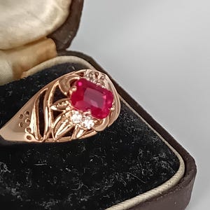 Pode incluir: Um anel em ouro rosa com uma grande pedra vermelha retangular como peça central. O anel é cravejado com pedras transparentes menores e apresenta um design ornamentado e vazado. É apresentado numa caixa de joias castanha vintage com interior em veludo preto.