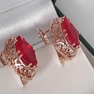 May include: Rose gold cufflinks with a filigree design and large, teardrop-shaped red gemstones. The cufflinks are displayed in a white box, showcasing their intricate details and vibrant color.