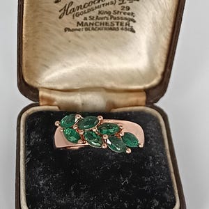 May include: A rose gold ring with a cluster of emerald-cut green gemstones. The ring is displayed in a vintage brown jewelry box with the text "Hancock & Son" and "Diamond Merchants".