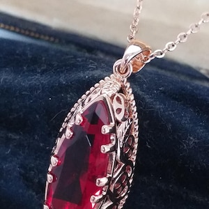 May include: Rose gold pendant necklace featuring a large, faceted, rectangular red gemstone. The gemstone is set within an ornate, filigree-style frame. The necklace chain is delicate and also rose gold.