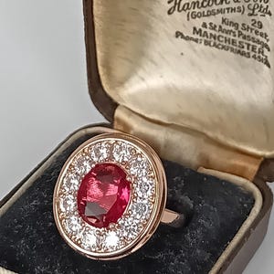 Puede incluir: Un anillo de oro rosa con una gran piedra roja de corte ovalado rodeada por un halo de pequeñas piedras transparentes. El anillo se presenta en un joyero forrado de terciopelo marrón con el texto "Diamond Merchants, Hancock & Son (Goldsmiths) Ltd".