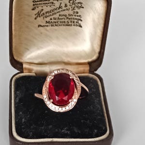 Puede incluir: Anillo de oro rosa con una gran gema roja ovalada, rodeada por un halo de piedras claras más pequeñas. El anillo se muestra en un joyero marrón vintage con letras doradas.
